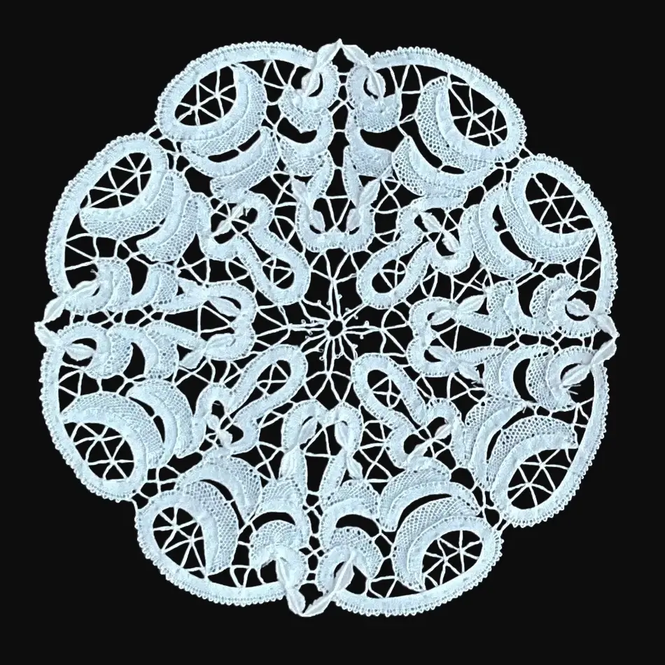 Ručno rađeni tabletić od čipke na batiće napravljen od tankog bijelog konca, u obliku kruga ispunjenog zavijutcima u obliku cvijeta s listićima, proporcionalno raspoređenih prema sredini čipke sa dodatnim malim elementima listića na površini čipke.