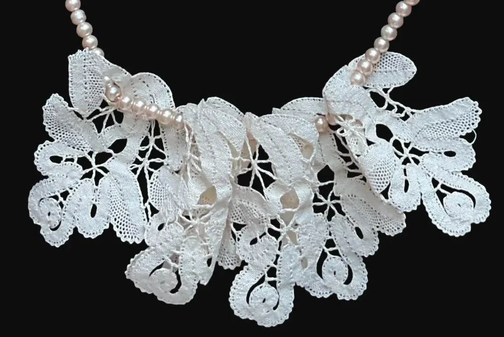 Ručno izrađena čipkana kragna u obliku zavijutaka, izrađena od tankog krem konca koja je spojena sa kratkom ogrlicom od blago rozih okruglih umjetnih bisera u jednu cjelinu.