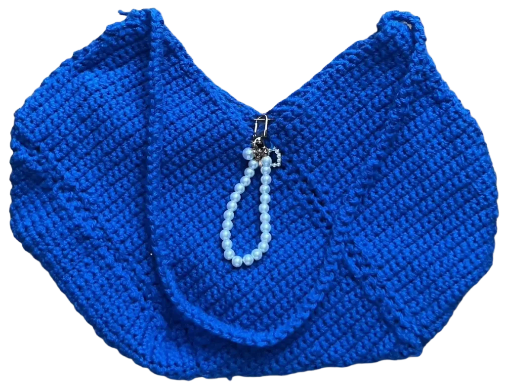 Ručno rađena velika heklana torba snažne plave boje sa drugom heklanom ručkom i ukrasnom bisernom kopčom koja visi na sredini torbe.